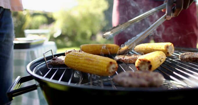 Grilling corn and burgers, man using tongs at outdoor barbecue gathering
