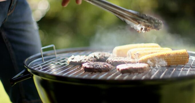 Grilling burgers and corn on barbecue grill, african american man using tongs for cooking, outdoors