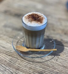 Close-up of a freshly made cappuccino served in a glass cup with creamy foam and cocoa powder, on a wooden table with a glass saucer and wooden spoon.