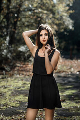 A girl with long dark hair standing against the background of a forest. She is wearing a sleeveless black dress
