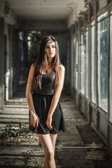 young woman with long dark hair standing in an abandoned building. She is wearing a short black dress and black high heels. The building looks old and abandoned