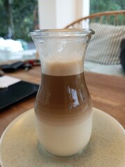 A close-up of a beautifully layered latte served in a transparent glass cup on a ceramic saucer, showcasing the separation of milk and coffee with foam on top.