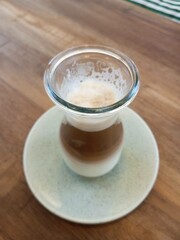 A close-up of a beautifully layered latte served in a transparent glass cup on a ceramic saucer, showcasing the separation of milk and coffee with foam on top.