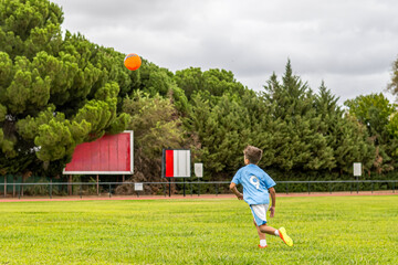 Young boy with the number 9 on his back is running on a soccer field, chasing an orange ball,...