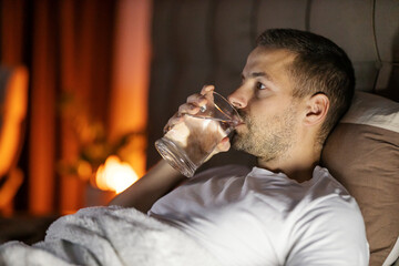 Side view of man lying in bed and refreshing himself with glass of water at night.