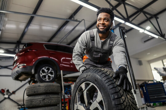 Portrait of smiling african american auto mechanic rolling and preparing tire for changing at car service.