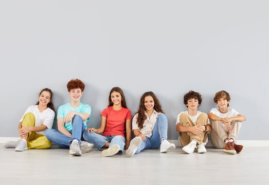 Group of happy students or friends sits together on the floor for a fun portrait on a grey background. The kids are enjoying time as a team, friendship and unity in this diverse and lively setting.