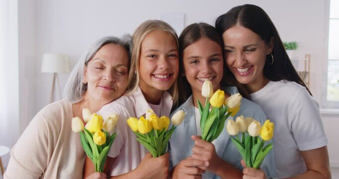 Joyful family two daughters, mother and grandmother sharing warm moment, smiling and embracing while holding flower bouquets of yellow and white tulips, celebrating International Women Day together.