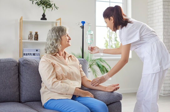 Friendly young female medical nurse helping senior elderly woman patient sitting on sofa in nursing home in receiving intravenous IV drip infusion and vitamin injection therapy to boost immune system.