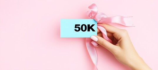 Fototapeta premium Female Hand Holds Card Saying 50K with Beautiful Pastel Pink Ribbon, Feminine Celebration Concept