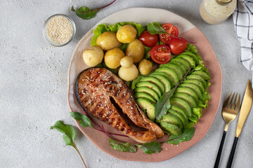 Baked salmon steak sprinkled with sesame seeds served with avocado, cherry tomatoes and potatoes on plate on light gray background