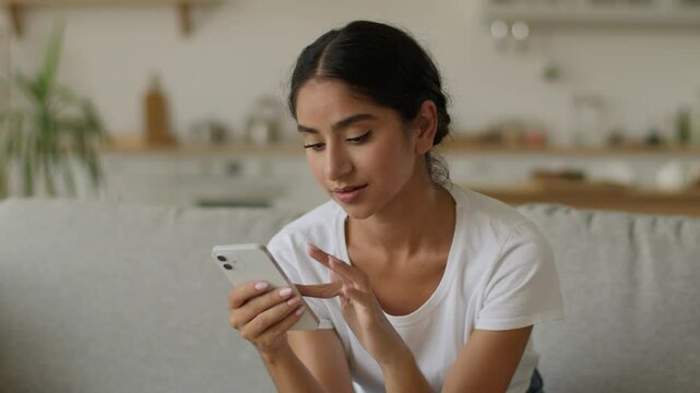 Indoors homeowner Indian woman Arabian female ethnic girl at home Hindu lady smiling looking at mobile phone using smartphone holding gadget addict browsing social media scrolling app shopping online