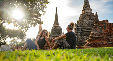 Two women enjoying a sunny afternoon at Ayutthaya Historical Park in Thailand, sitting on grass near ancient temples and stupas, surrounded by nature and cultural heritage
