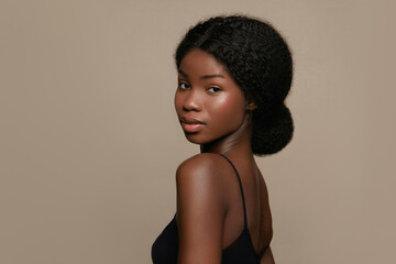 Elegant African American woman with natural textured hair styled in a low bun with a clean glowing skin, wearing a black sleeveless top, looking over her shoulder against a beige background