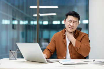 Fototapeta premium Asian sick man at workplace has sore throat, severe sore throat pain. Man with cold working inside office at workplace.