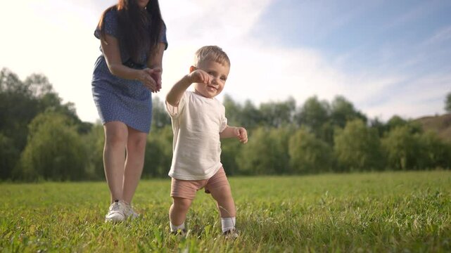 A mom is walking her baby while learning to walk. family holding green concept. A baby is taking the first steps in a park. Mom and baby learn to walk in park. family lifestyle motherhood concept.