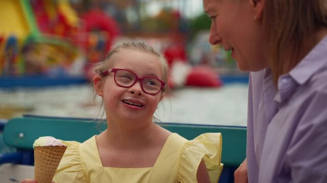 Smiling child with Down syndrome enjoys ice cream while engaging in conversation with her mother. They are seated near water, surrounded by vibrant attractions. Concept inclusion, and family bond