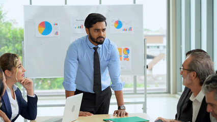 stressed young indian business manager standing and presenting to multiracial group of diversity colleagues in front of white board in meeting at boardroom in office . leader