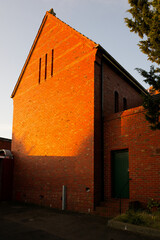 Rear of a red brick church building illuminated by the setting sun, with paved courtyard and green wooden door in the shade