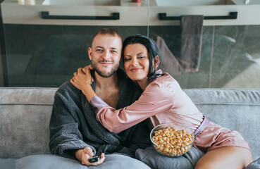 Cozy on the couch with popcorn and TV remote, a couple shares affectionate moments draped in robes,...