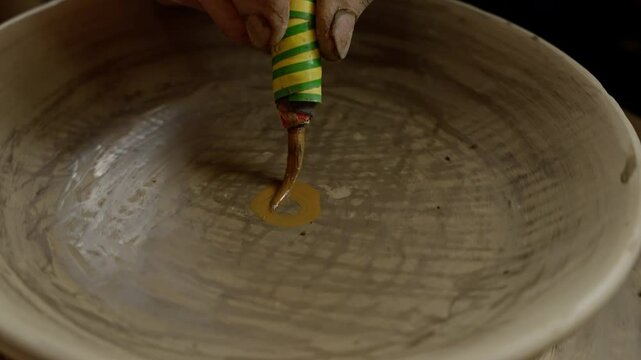 Skilled craftsman carefully painting intricate spiral design on ceramic bowl using thin brush and colored clay slip, capturing delicate artistic process in slow motion