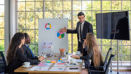 Collaboration is a key to best results. Group of young modern people in smart clothing wear planning business strategy while young man pointing at infographic displayed on the glass wall in the office