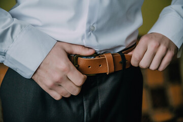 A man is wearing a brown belt and adjusting it. Concept of formality and attention to detail, as the man is carefully adjusting his belt to ensure it is properly fastened