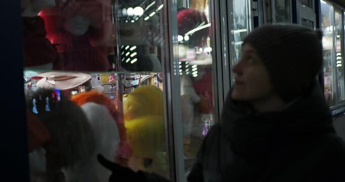 Retail, night city. A young woman in winter clothes smiles looking at the illuminated window of a small store. Hand held. Siberia.