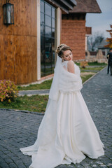 Fototapeta premium A bride is standing in front of a building, wearing a white dress and a fur stole. She is smiling and looking towards the camera