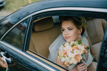 A woman in a wedding dress is sitting in a car with a bouquet of flowers. The car is black and has a shiny interior. The woman is happy and excited about her upcoming wedding