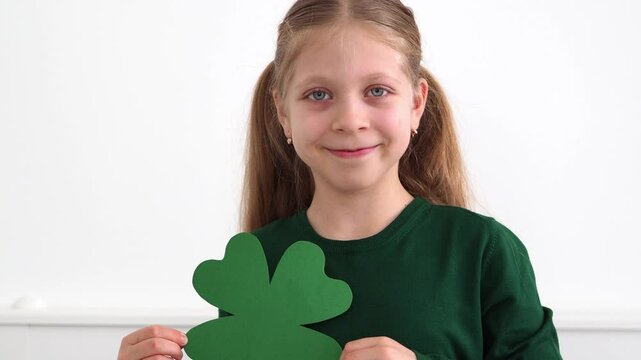 Hold a large green clover trefoil in your hand. A happy child in a St. Patrick's Day outfit on a light background. The symbol of St. Patrick's Day