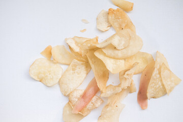 Scattered cassava chips (keripik singkong) on a white surface. The chips are thin, crispy, and have a light golden-brown color with some slightly darker or reddish tinges.