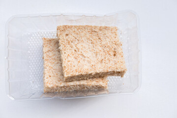 Two slices of whole wheat bread in a clear plastic container on a white background. The bread has a visible texture with small air pockets and bran flecks.