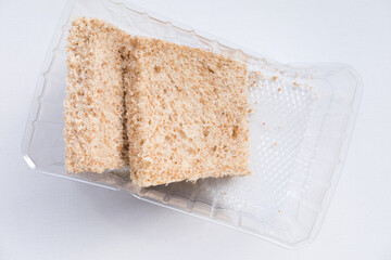 Two slices of whole wheat bread in a clear plastic container on a white background. The bread has a visible texture with small air pockets and bran flecks.