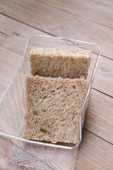 Two slices of whole wheat bread in a clear plastic container on a white background. The bread has a visible texture with small air pockets and bran flecks.