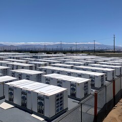 A largescale outdoor array of white lithiumion battery storage units, enclosed by a fence, under a clear blue sky. The installation occupies a substantial area.