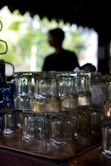 Stack of clear glass cups, arranged upside down, on wooden surface, with blurred greenery and dark silhouette in background. 