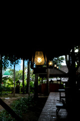 A black kerosene lantern hangs lit against a blurred green and brown background of trees and...