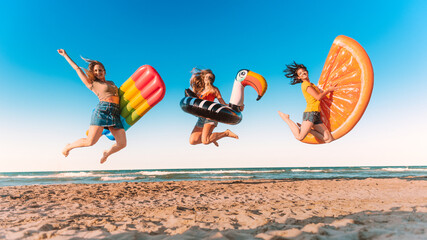 Three happy girls on jumping motion with inflatable mattresses at the beach for celebrating summer vacation - Friends women having fun outdoors on seaside holidays - Joy, carefree and party concept © charmedlightph