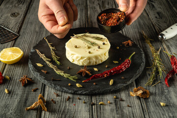 Hands sprinkle aromatic herbs and spices over a creamy cheese wheel, surrounded by vibrant dried...