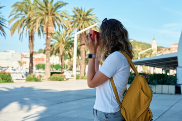 Female traveler in casual clothing taking picture in coastal town. Woman using orange retro instant...