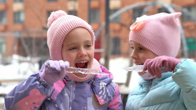 Two happy little girls fun play in snowy yard and want to bite icicles in winter outdoors. Children in winter