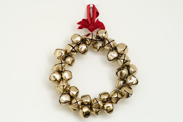 Golden jingle bell wreath with red ribbon hanging on white wall. Minimal holiday decoration. Christmas celebration and winter season concept for design and print. Studio shot.