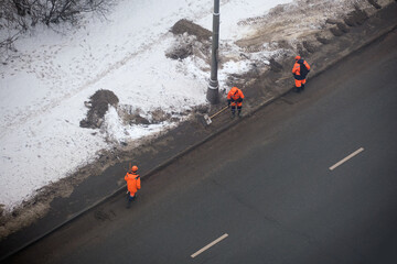 Workers in bright orange uniforms clearing snow and dirt along a roadside in winter conditions....