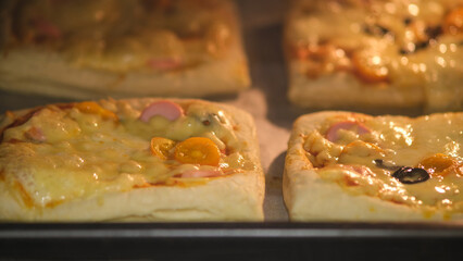 Homemade square pizzas with melted cheese, cherry tomatoes, olives, and sausage slices baking in the oven. Close-up food photography of freshly baked pizza slices.