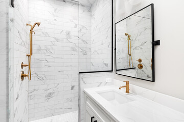 A white bathroom with a marble countertop, gold faucets, black mirror, and a shower with marble...