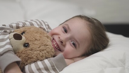 Child smiles happily and hugs plush teddy bear tightly lying on pillow in bedroom. Daughter preschooler wants mother to tell fairytale before falling asleep