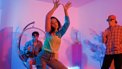 Fototapeta premium Attractive happy dancer looking at camera while smart woman moving at rhyme with neon light. Professional hispanic performer break dancing while wearing colorful cloth with diverse friend. Regalement.