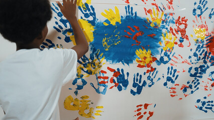 Obraz premium Back view of happy african boy painted the stained wall with colorful hand while wearing casual white shirt in art lesson.Smart student use hand print to make creative artwork. Education. Edification.