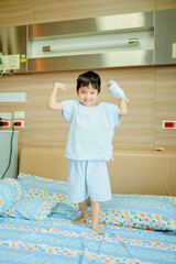 Young boy wearing light blue hospital pajamas, standing on a bed with a cheerful expression